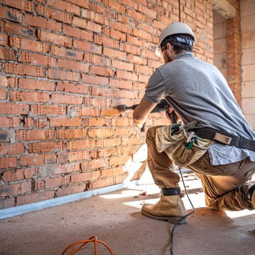 A builder works with a drill at a construction site. Handyman at a construction site in the process of drilling a wall with a perforator.